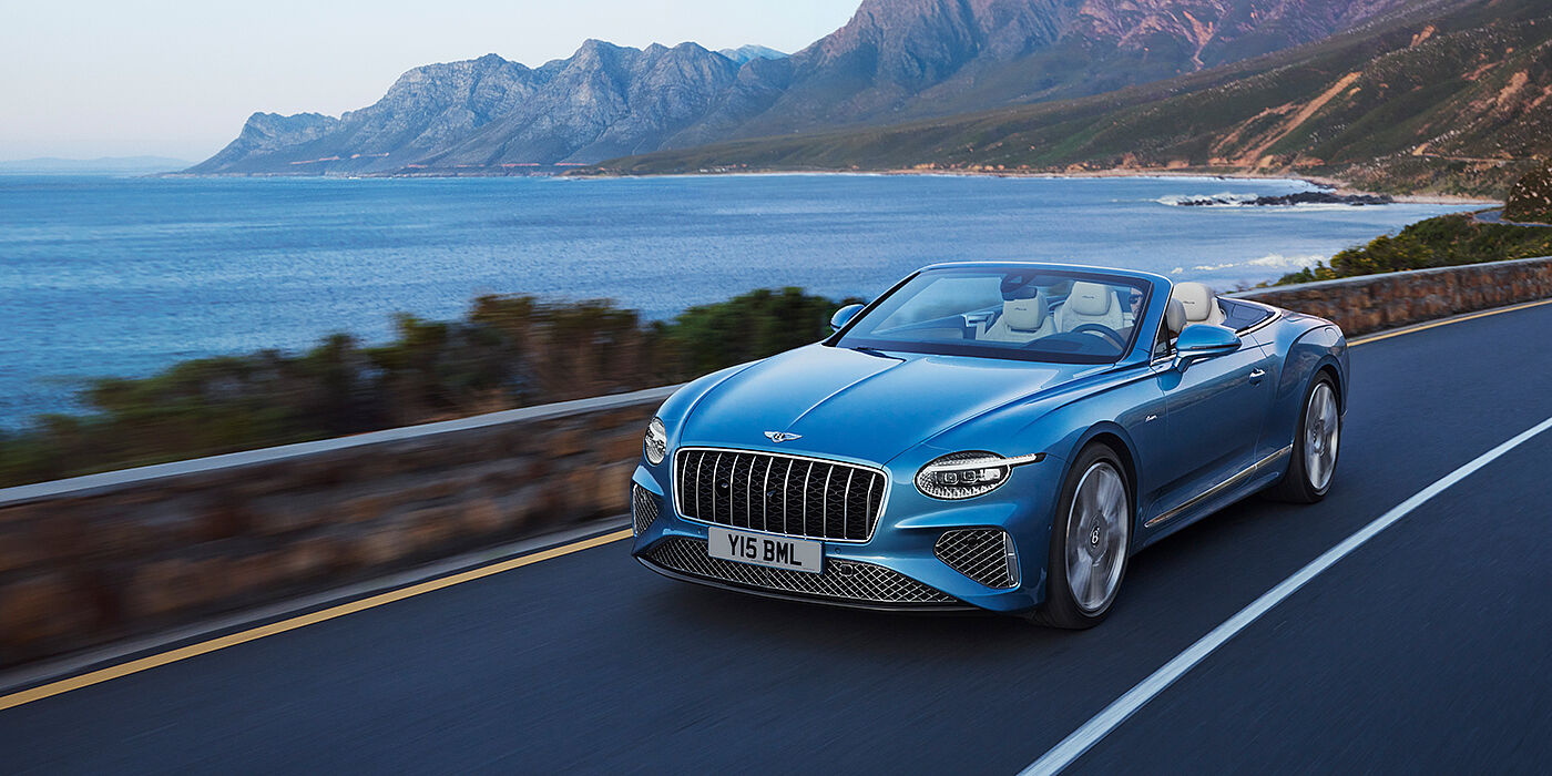 Bentley Manchester New Bentley Continental GTC Azure convertible in Light Windsor blue paint with 22 inch Azure wheels and chrome grill, driving dynamically by the sea