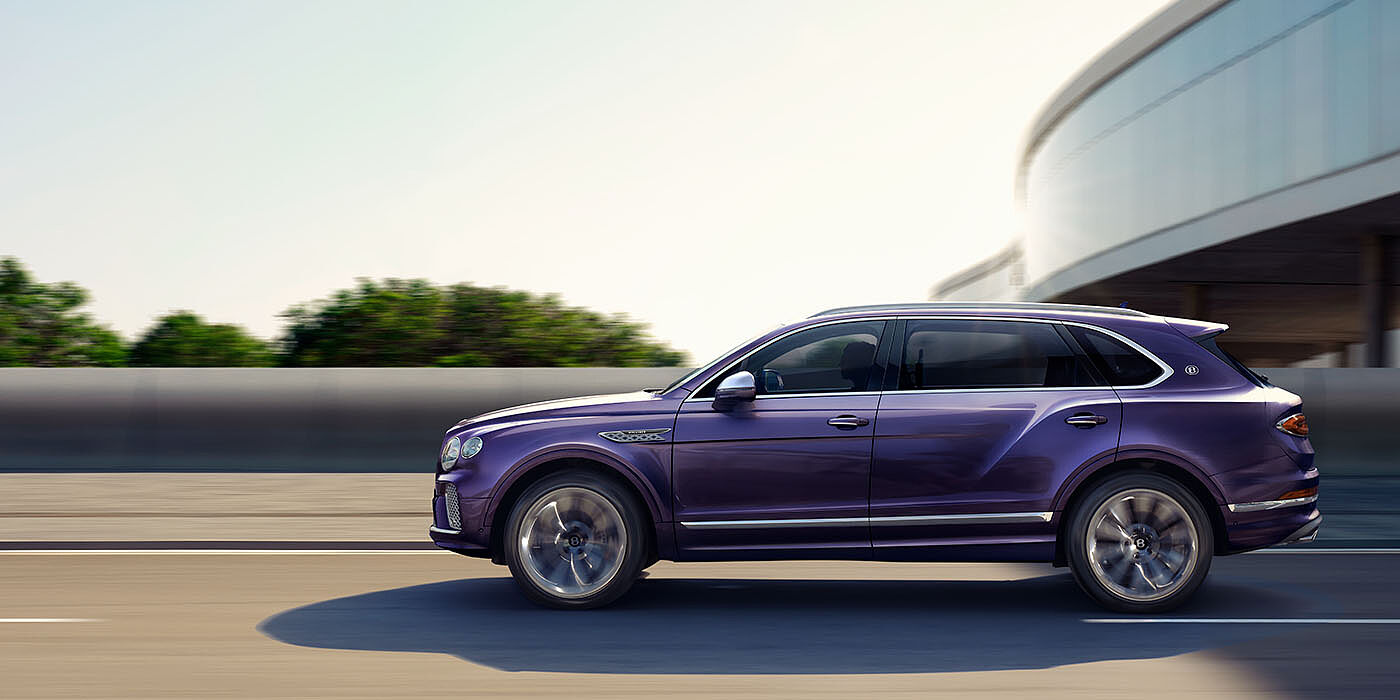 Bentley Manchester Bentley Bentayga Extended Wheelbase Mulliner luxury SUV profile in Tanzanite Purple paint, driving on an country road past trees.