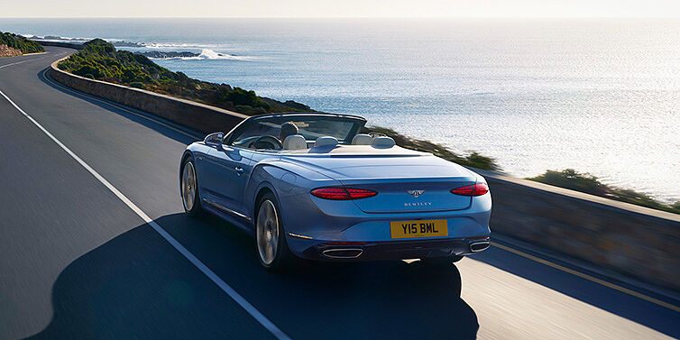 Bentley Manchester New Bentley Continental GTC Azure convertible rear view in Light Windsor blue paint with 22 inch ten spoke wheels driving dynamically on an ocean road in the sunshine